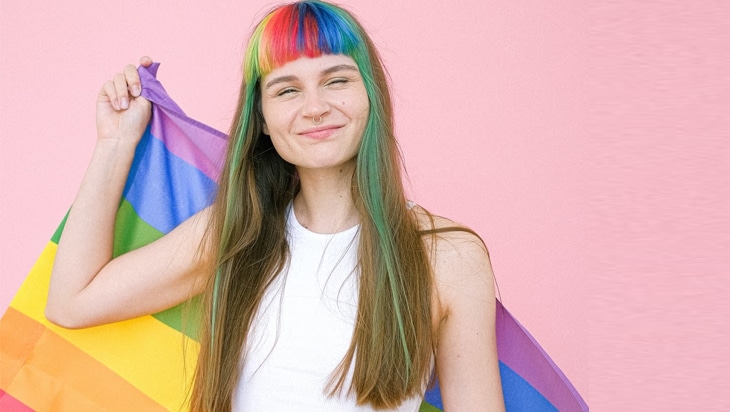 Queer person wearing a white top and coloured hair highlights holding the Pride flag