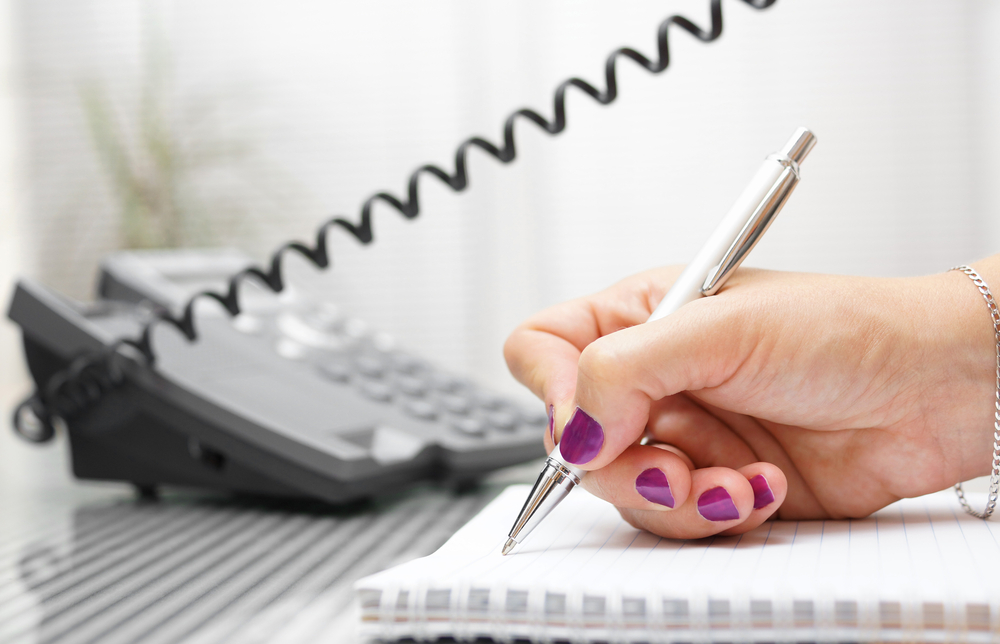Close-up shot of a telephone and hands taking notes with pen and notebook.
