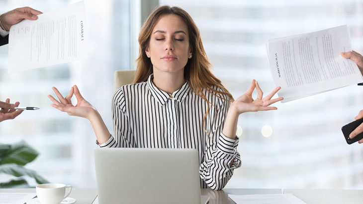 A female employee meditating in her office surrounded by hands holding paperwork.