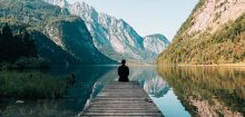 A man meditating on the edge of a wooden dock as he looks at a picturesque lake.