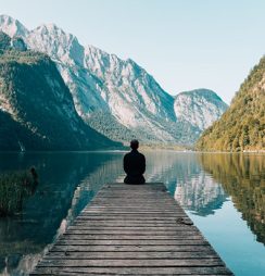 A man meditating on the edge of a wooden dock as he looks at a picturesque lake.