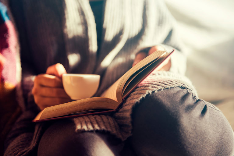 A woman holding a cup of coffee while reading a book about addiction recovery