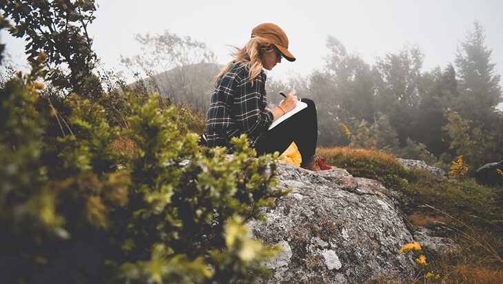 A woman sitting on a rock while she writes on her journal