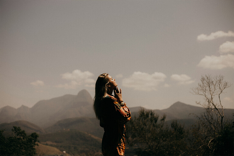 A woman standing in a desert with her hands clasped and face turned toward the sky