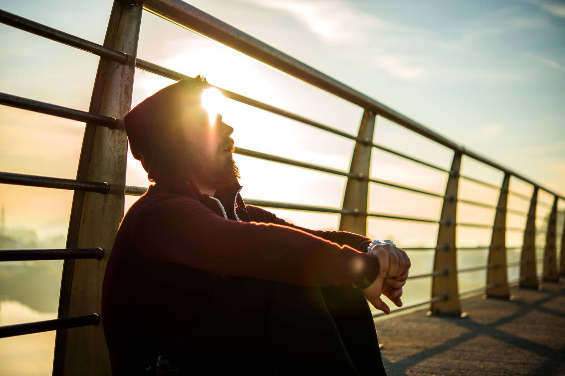 A hooded man sitting on a bridge leaning against the railing while the sun rises in the background