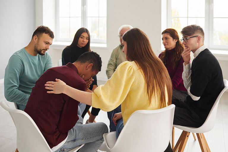 Group therapy session with a woman in a yellow shirt comforting the man next to her