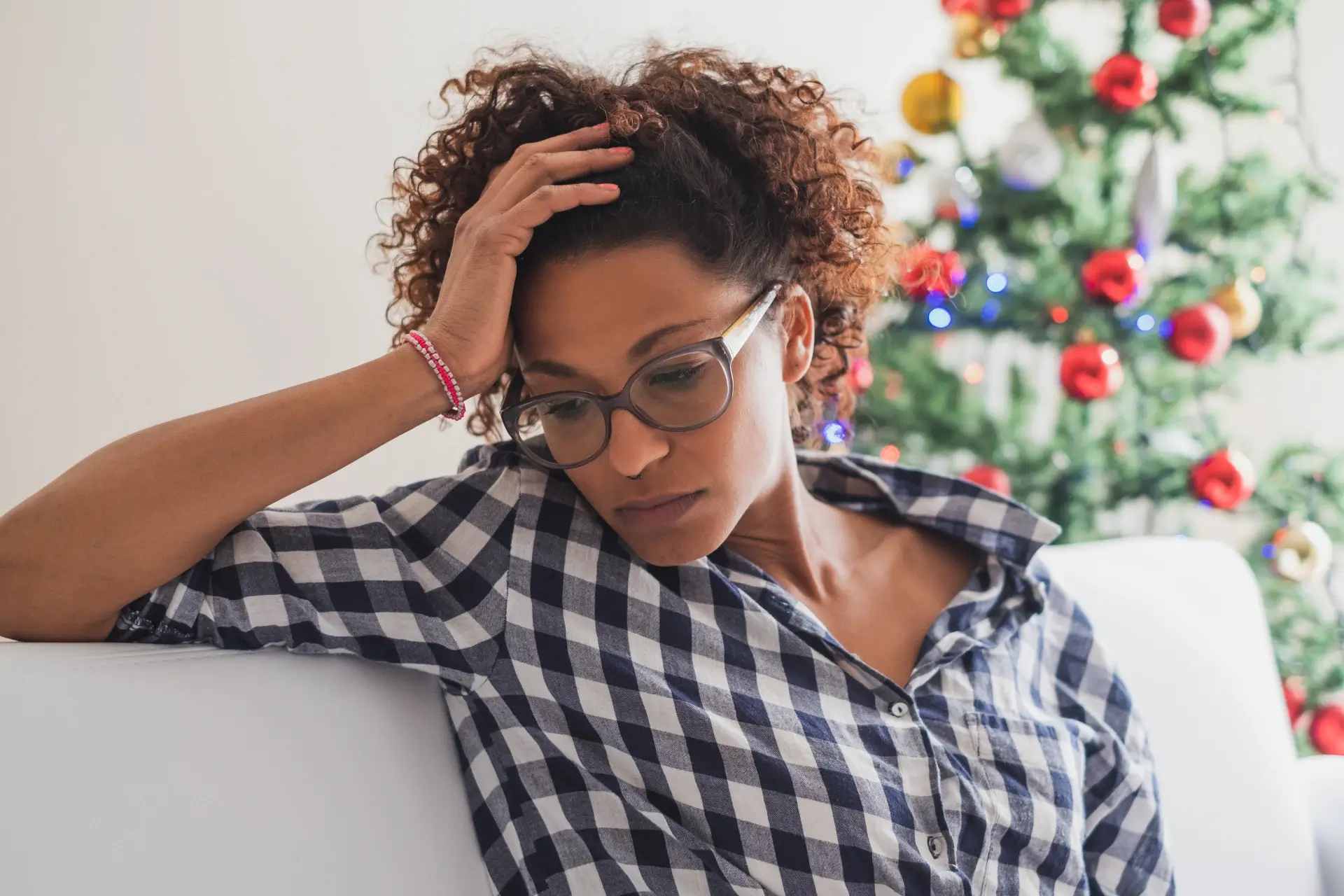 A woman sits on a couch in front of a Christmas tree with her head in her hand