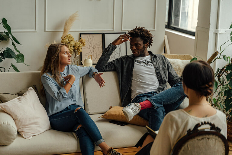 A man and a woman sit on a couch opposite a therapist in a couples therapy session. 