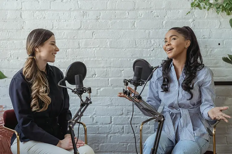 Two women sit in front of microphones while recording a podcast.