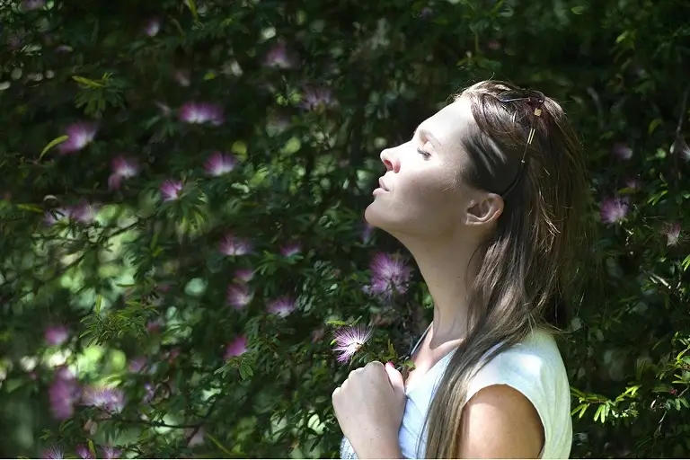 A woman standing amidst greenery and purple flowers with her eyes closed, taking in the smell and feeling the sunlight.