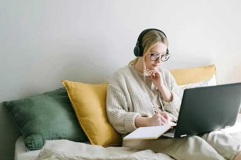 A woman wearing headphones sits in bed while journaling in a notebook in front of her laptop.