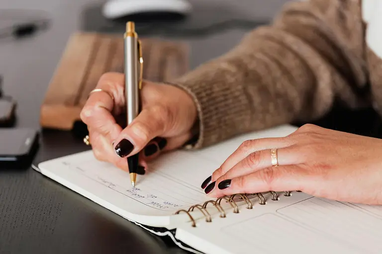 Close-up of a woman's hands writing in a spiral notebook with a gold pen.