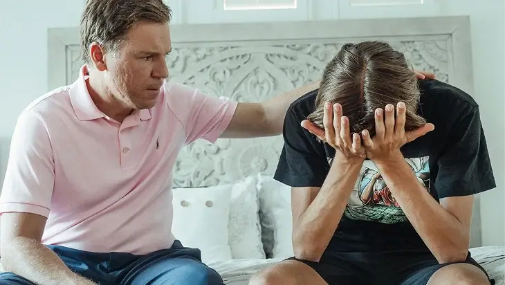Father comforting distressed son sitting on bed with head in hands.