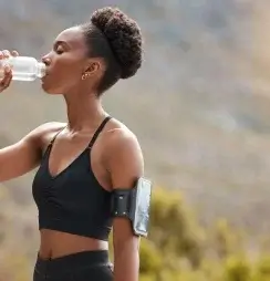 While jogging through nature, a woman pauses to take a refreshing sip from her water bottle, reflecting on summer self-care ideas.