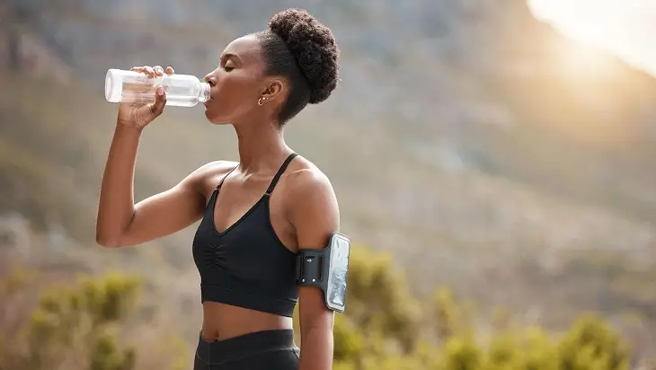 While jogging through nature, a woman pauses to take a refreshing sip from her water bottle, reflecting on summer self-care ideas.