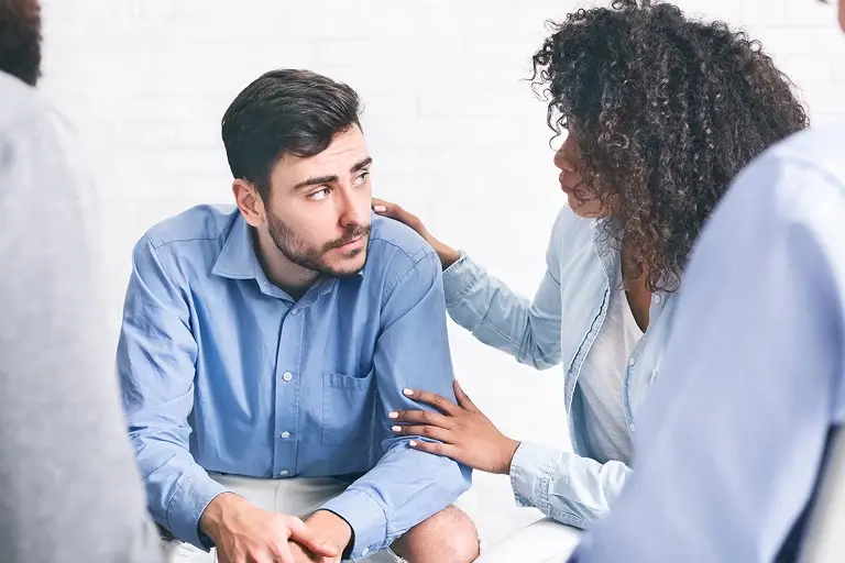 A woman comforts a man in a group setting.
