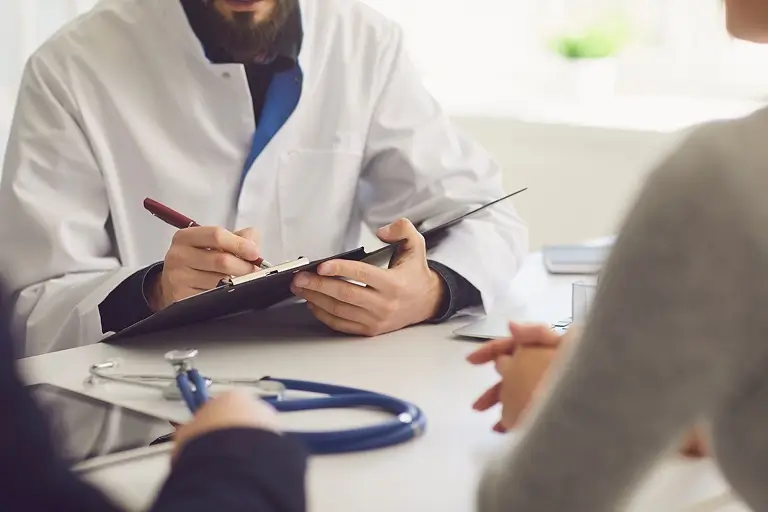 A patient and advocate sit in front of a physician with a clipboard.